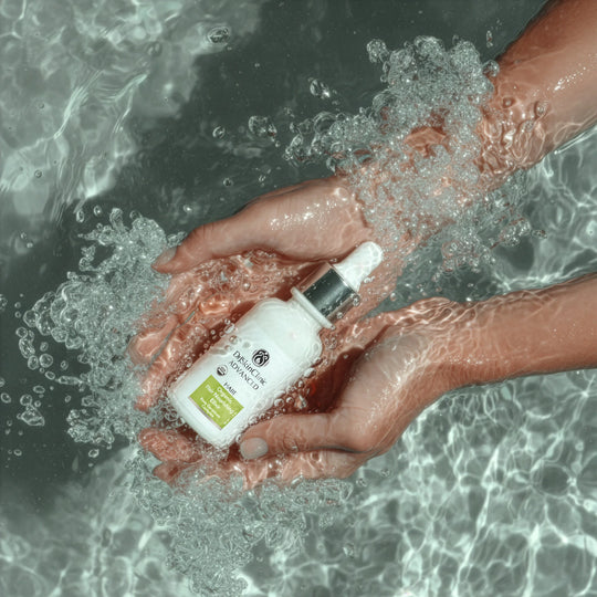 Hands holding DrSkinClinic Advanced Organic Hair Nourishing Elixir dropper bottle in water, highlighting hydration and scalp care theme